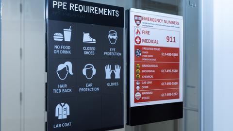 A sign displaying PPE requirements for a lab, including no food or drink. Next to it is an emergency numbers sign listing contacts.